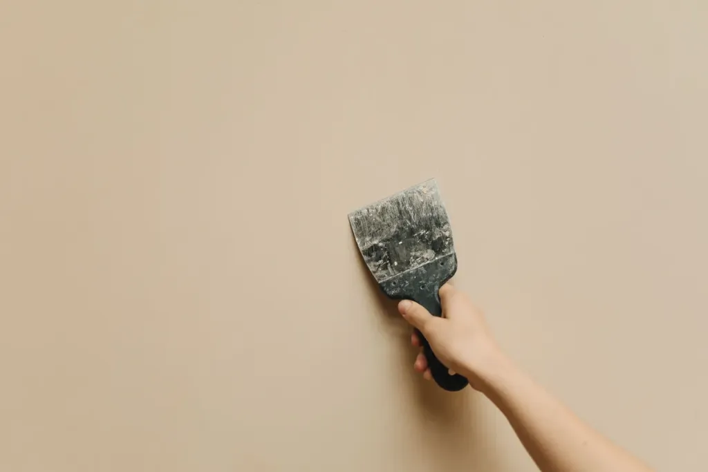 Hand houdt een plamuurmes tegen een kale beige muur tijdens het opknappen van de wand
