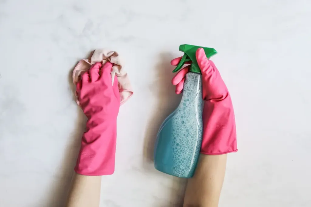 Handen in roze handschoenen maken een wit oppervlak schoon met een sprayflacon en doek